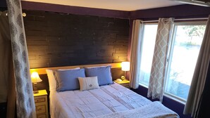 Uma cama bem arrumada com edredom branco e duas almofadas, ladeada por mesas de cabeceira com abajures, em um quarto com parede de tijolos escuros e janela com cortinas.