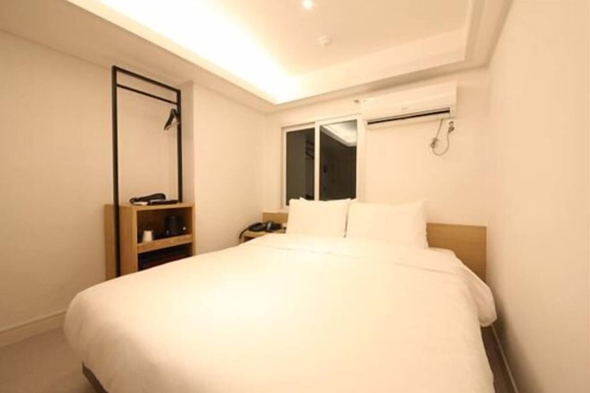 A hotel room with a bed, a nightstand, a shelf, a window, and a ceiling light.