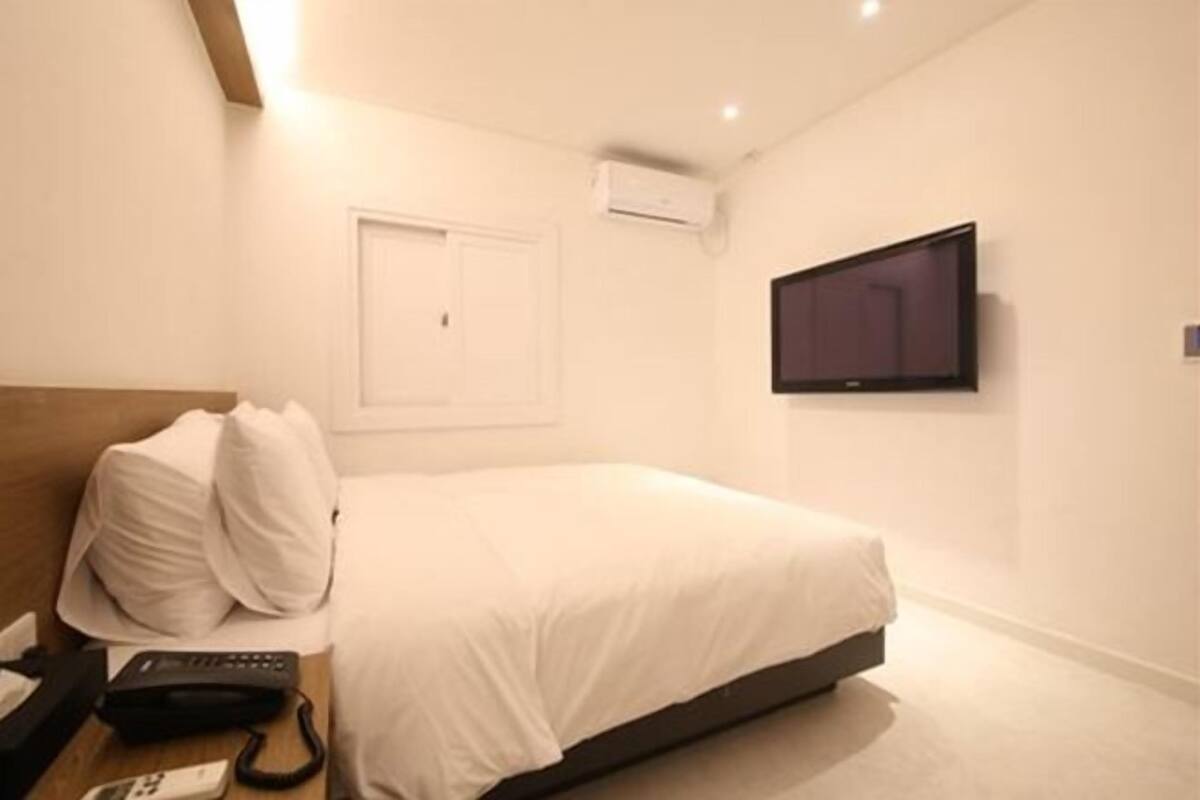 A hotel room with a bed, a TV mounted on the wall, a bedside table with a phone, and a door with an air conditioning unit.