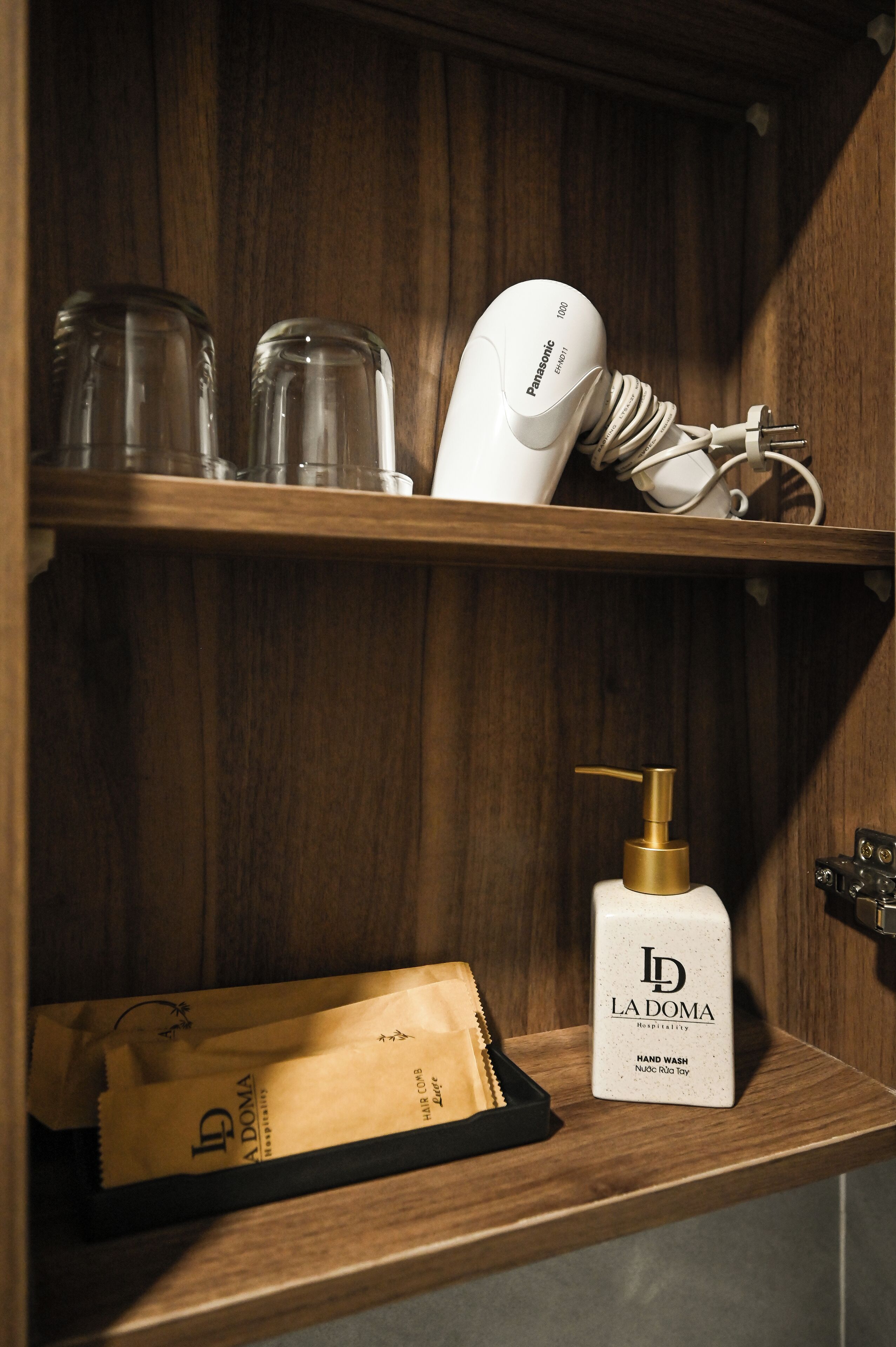 Une étagère en bois sur laquelle se trouvent un sèche-cheveux Panasonic, un distributeur de savon pour les mains de la marque La Doma et un sac contenant des produits de soins capillaires La Doma.