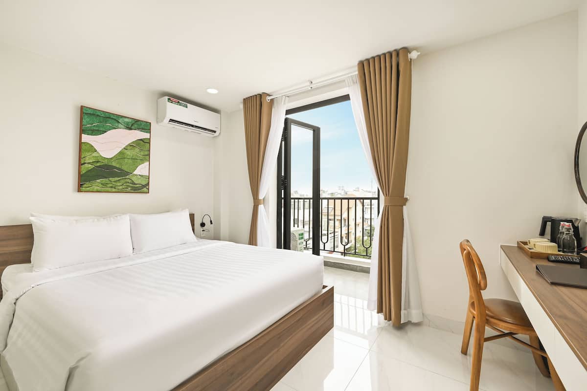 Una habitación de hotel con cama, escritorio, silla, ventana con cortinas, aire acondicionado y un cuadro en la pared.