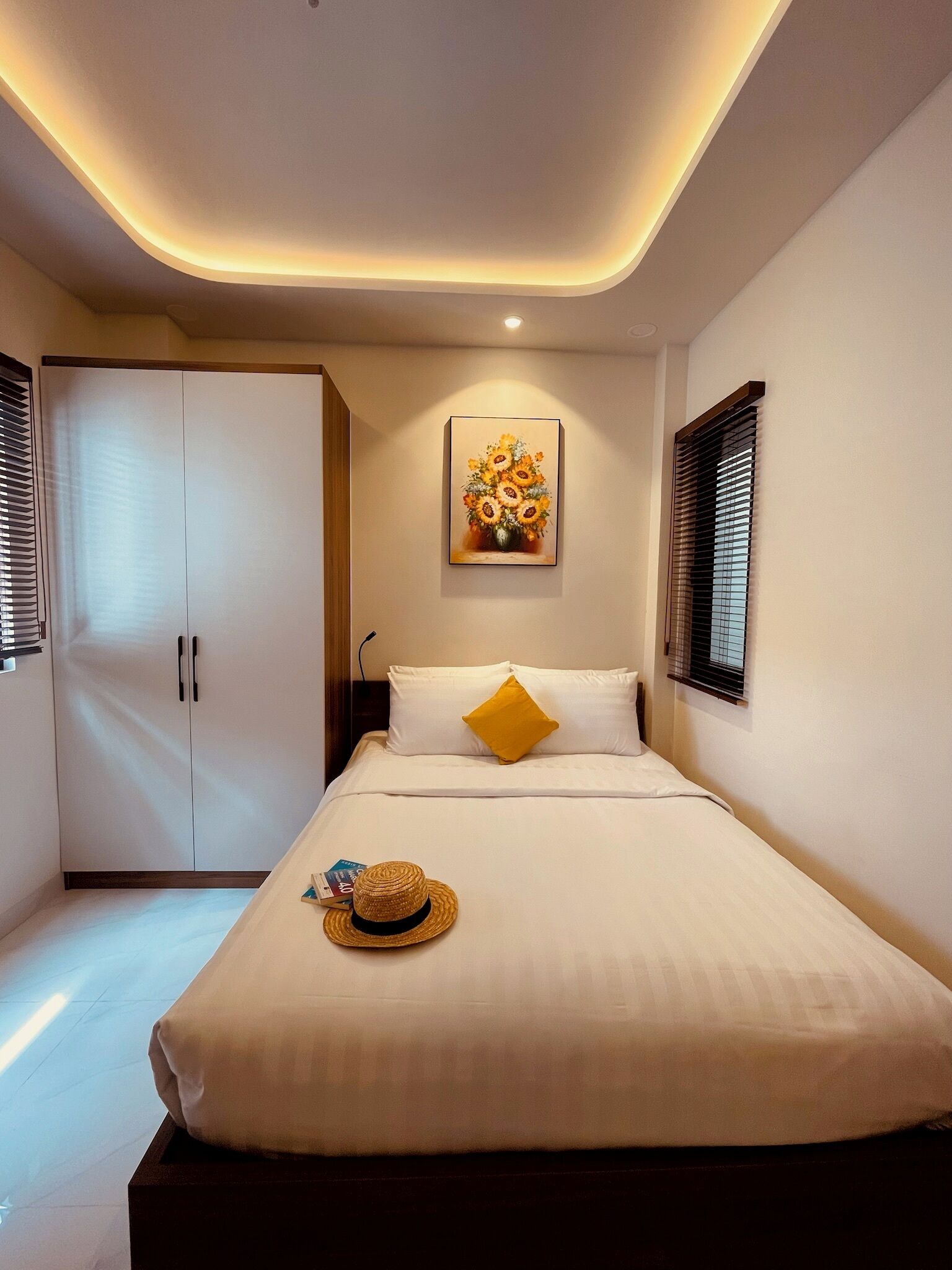 A neatly arranged hotel room with a bed, a wardrobe, a painting, and a window with blinds.