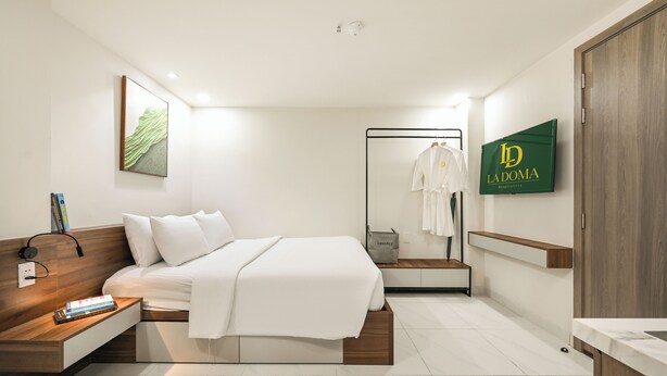 Une chambre d’hôtel moderne avec un lit, une table de chevet et un miroir. Un panneau vert portant l’inscription « La Doma » est visible sur le mur.