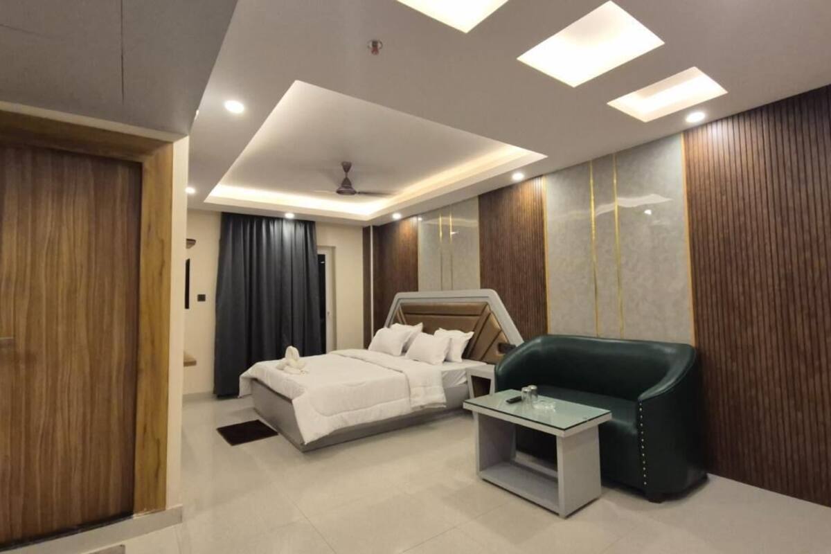 A modern hotel room with a bed, a glass table, a green armchair, and wooden paneling on the walls.