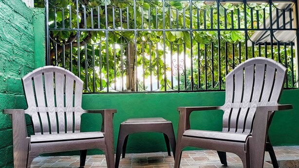 Two wooden chairs and a small table on a stone-tiled patio with a green wall and metal bars in the background.