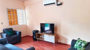 A living room with a flat-screen TV on a wooden TV stand, two sofas with green and black cushions, a large window with a decorative grille, and an air conditioning unit on the wall.
