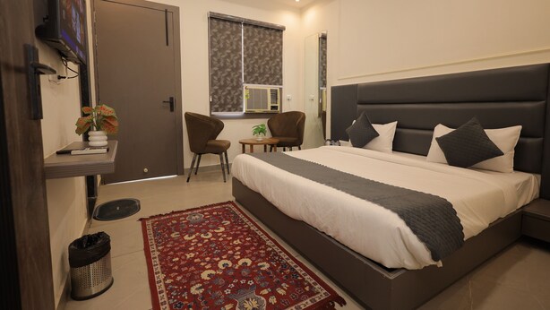 A bedroom with a large bed, a TV mounted on the wall, a window with blinds, a small table with a plant, and a chair. - HOTEL CRESTA INN (Agra)