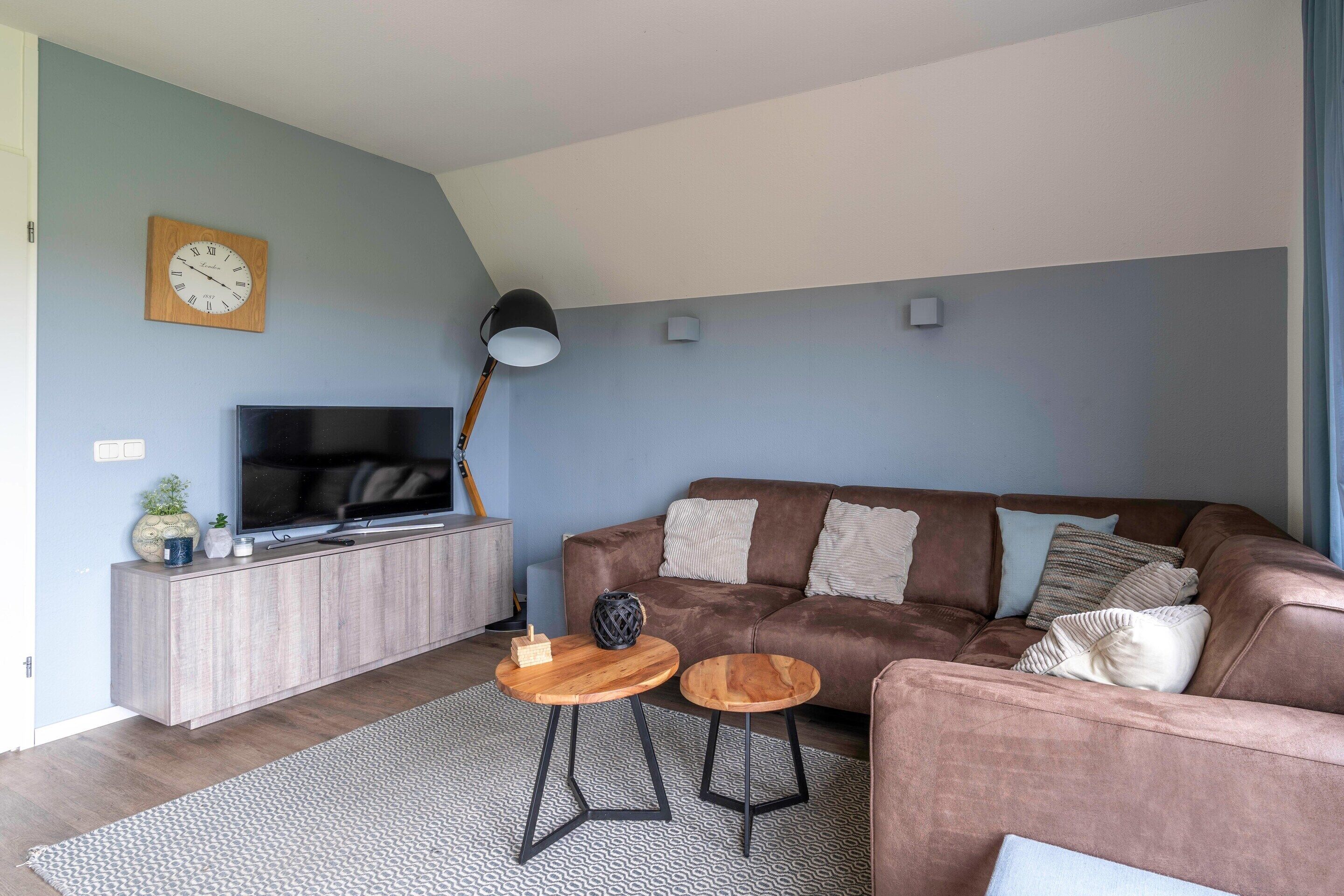 A modern living room with a brown leather sofa, a flat-screen TV, a wooden cabinet, and two small wooden tables.