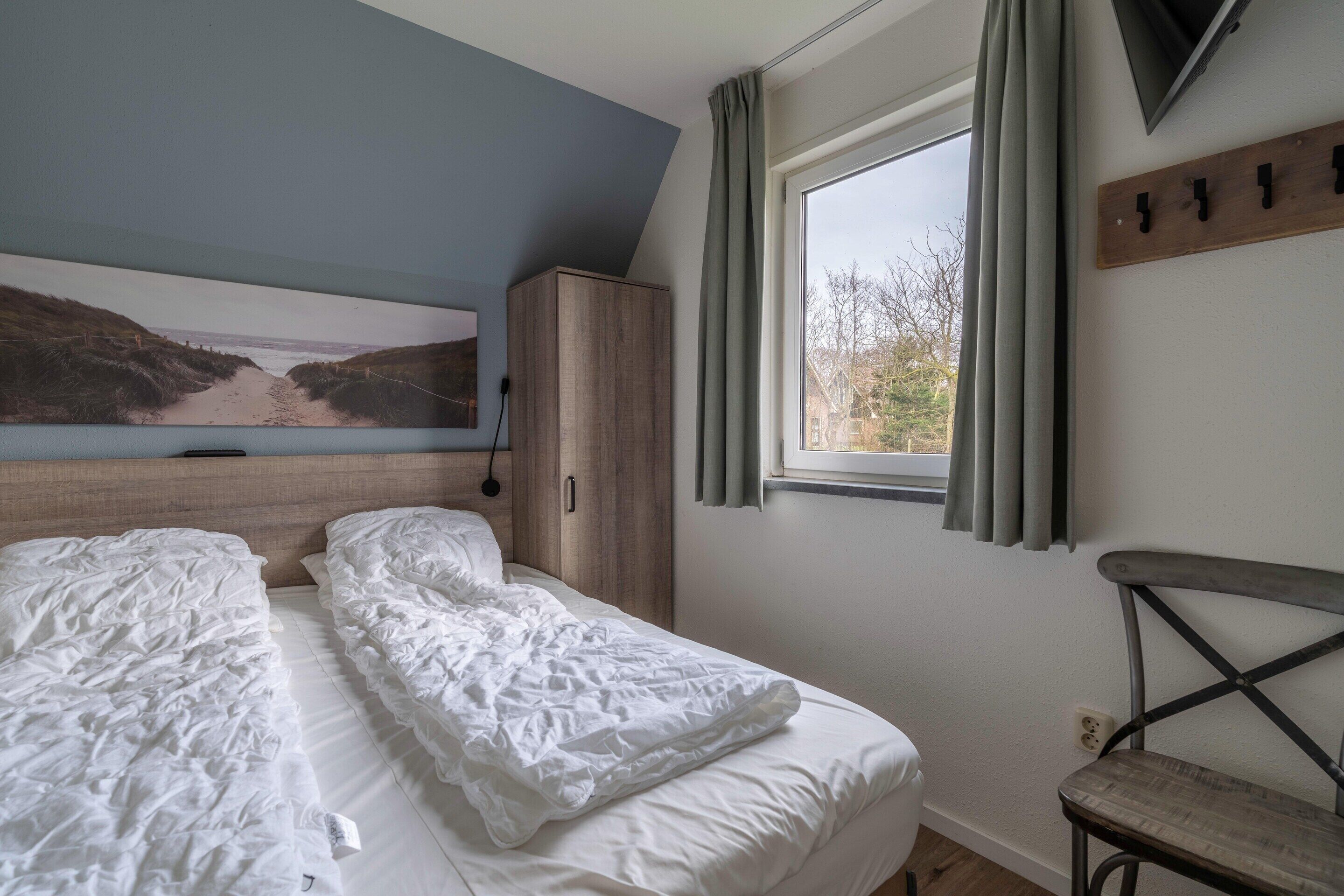 Ein Schlafzimmer mit einem Bett, einem Holzschrank, einem Fenster mit Vorhängen und einem an der Wand befestigten Fernseher.