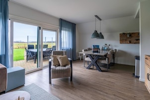 Uma sala de estar espaçosa com uma grande porta de vidro deslizante que dá para um pátio, uma mesa de jantar com cadeiras, uma poltrona confortável e uma luminária pendente.