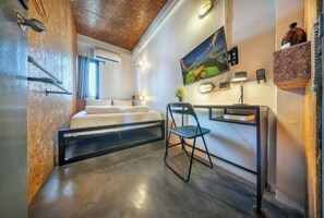 A modern hotel room with a bed, a desk with a chair, a TV, and a shelf with a bottle.