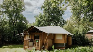 A small, rustic cabin with a porch and a covered outdoor area, surrounded by lush greenery. - Luxury Family Tent With Play Area (Balkbrug)