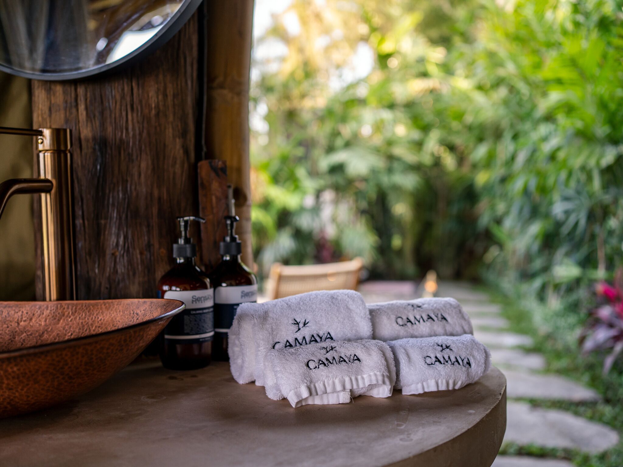 A bathroom with a copper basin, two bottles of soap, and three towels with "CAMAYA" printed on them.