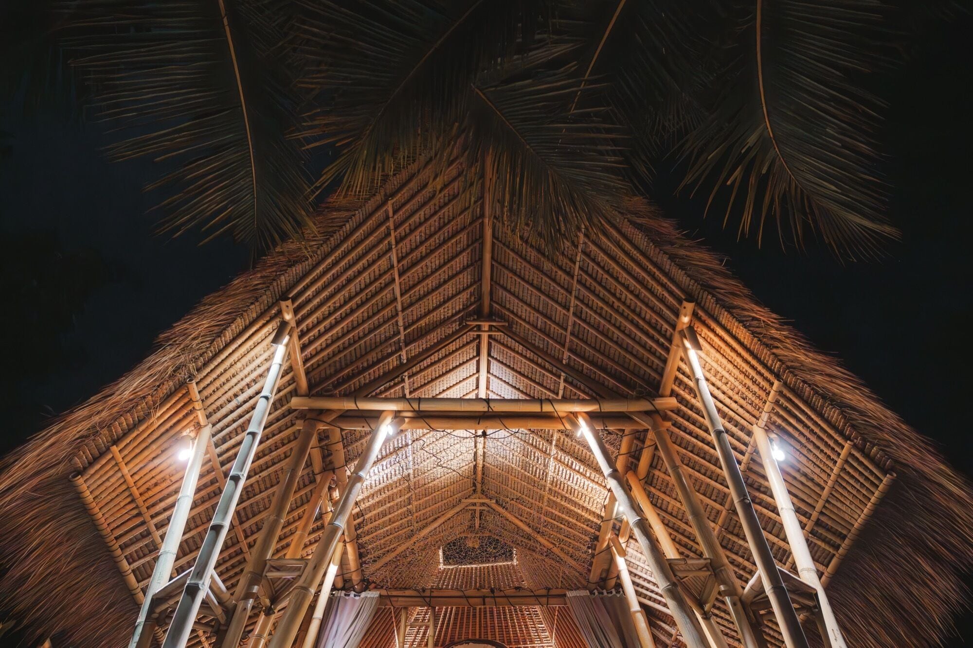 A thatched-roof structure with a central open space, possibly a communal area or entrance, surrounded by bamboo poles and supported by wooden beams.