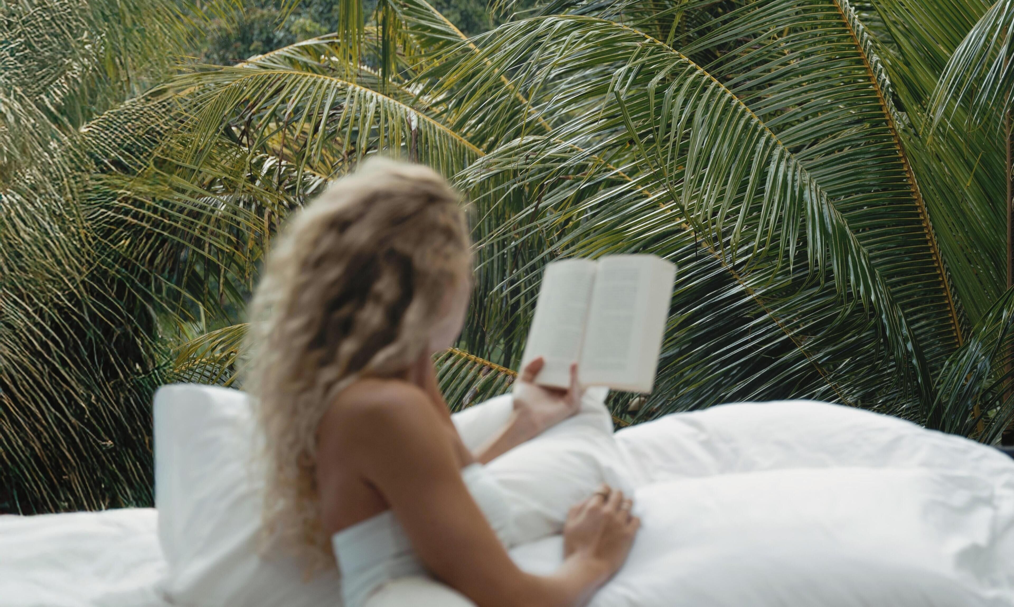 A person reading a book on a bed with white bedding, surrounded by palm trees.