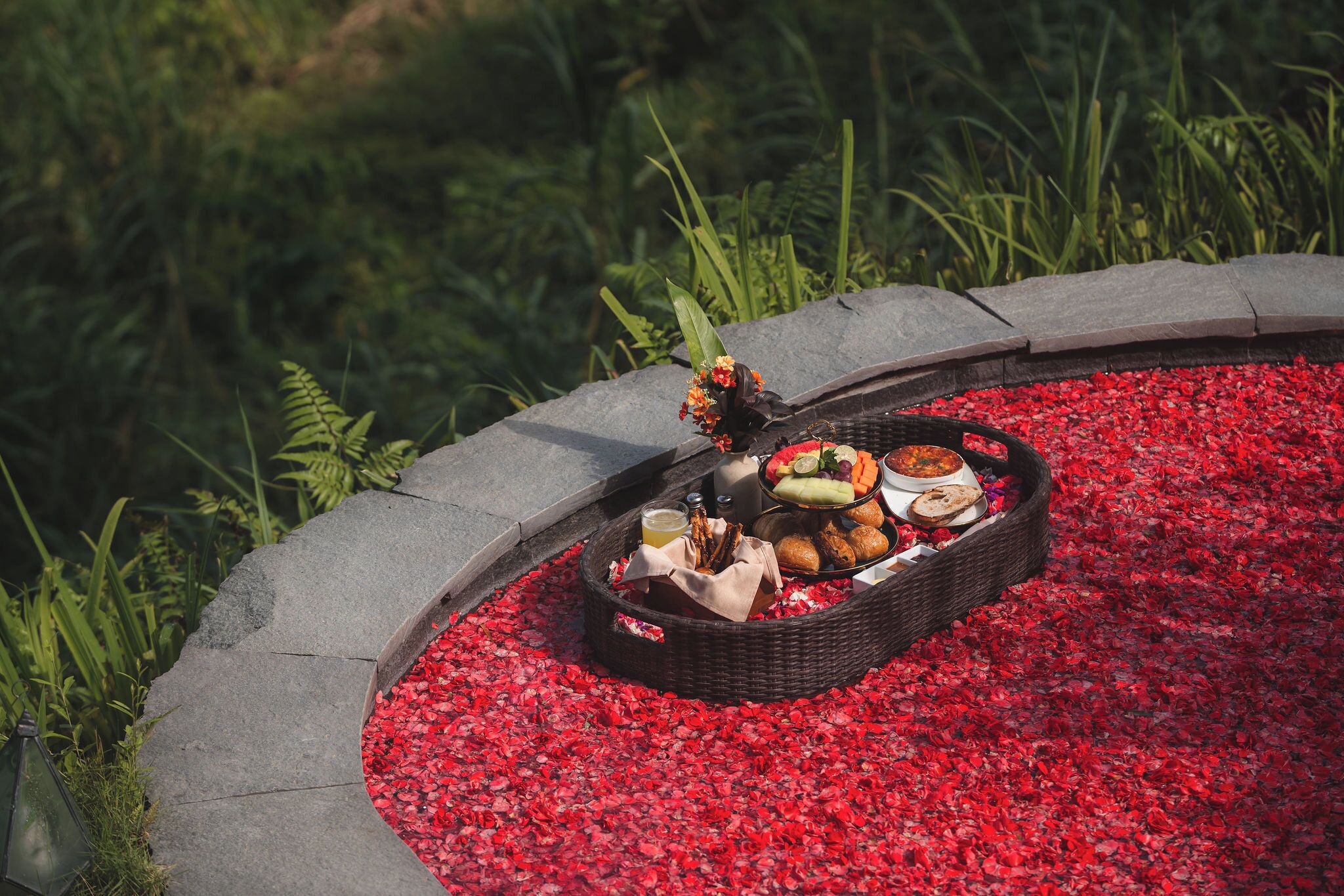 Un entorn de jardí amb un camí de pedra, un cistell decoratiu ple d'aliments i begudes, i pètals de flors vermelles al terra.
