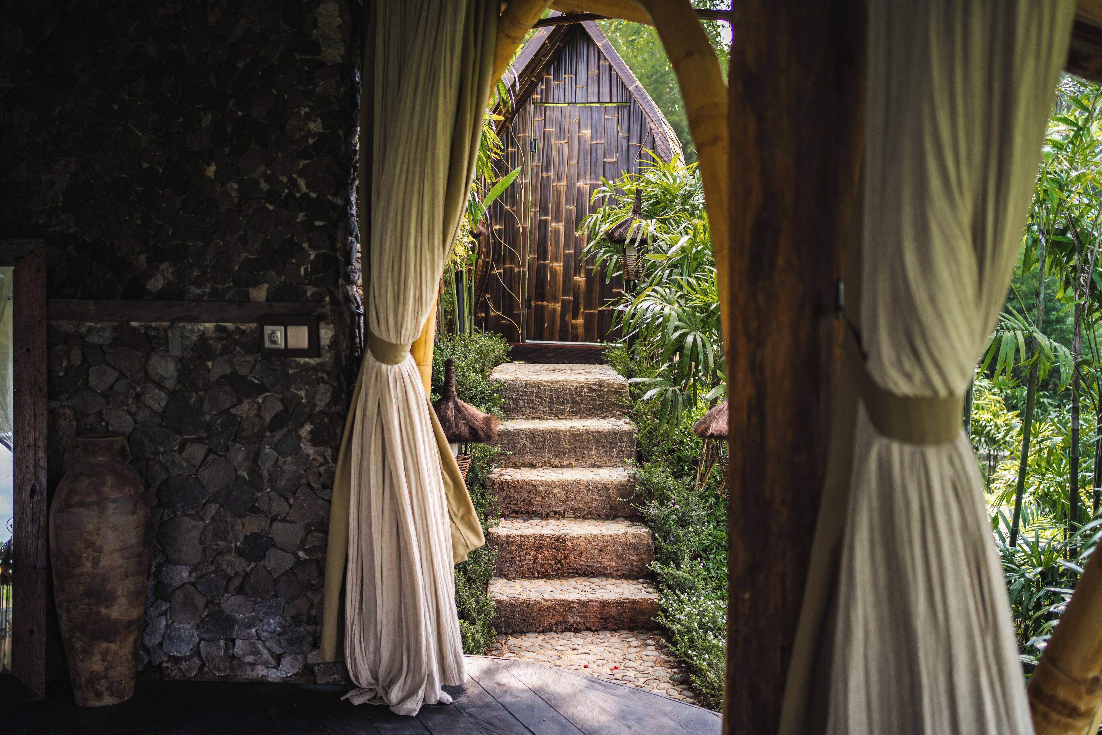 A stone wall with a wooden door, a staircase leading to a wooden structure, and a large vase.