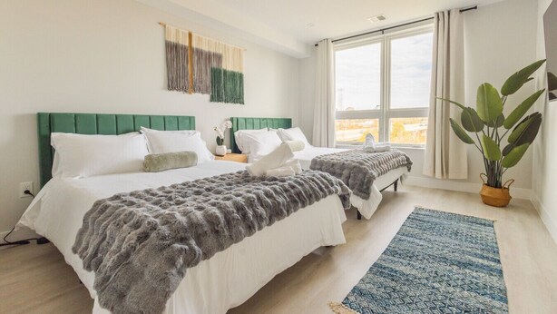 A hotel room with two beds, a large window, a green headboard, a plant, and a blue rug. - Spacious 3BR Loft | 15 From NYC & MetLife| Great Value | Groups Welcome (Jersey City)