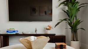 Una sala de estar moderna con una mesa redonda, un cuenco decorativo, una pequeña caja de madera, una planta en maceta, un televisor montado en la pared y una estantería con adornos y libros.