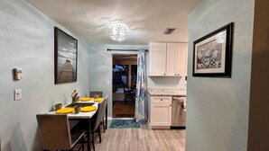 A narrow hallway with a dining area on the left, featuring a table with yellow plates and a framed picture above it. The kitchen is visible on the right with white cabinets and a countertop.