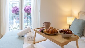 Habitación de hotel con una cama, una mesita de noche con una bandeja que contiene croissants, un bol de fruta y una taza.