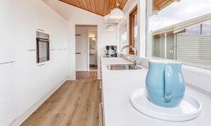 Een moderne keuken met witte kastjes, een houten vloer, een inbouwoven, een spoelbak en een raam met jaloezieën.