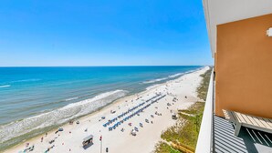 Blick auf einen Strand vom Balkon aus, mit blauem Himmel, Sandstrand, Sonnenschirmen und Liegestühlen sowie einem Rettungsturm.