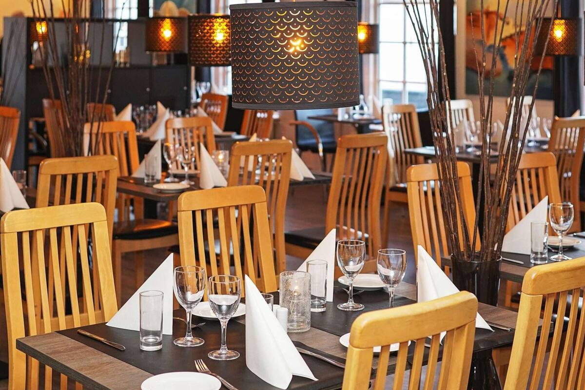Interno di un ristorante con sedie e tavoli di legno, apparecchiati con tovaglioli bianchi, bicchieri e posate.