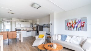 En moderne stue med sofa, sofabord og stole, et køkken i baggrunden og et farverigt billede på væggen.