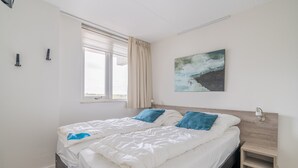 En dobbeltseng med hvidt og blåt sengetøj, et vindue med gardiner, et billede på væggen og et natbord.