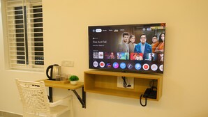A television mounted on a wall with a wooden shelf below displaying a coffee maker and a plant. - Aeroview Homestay Chennai Airport (Chennai)