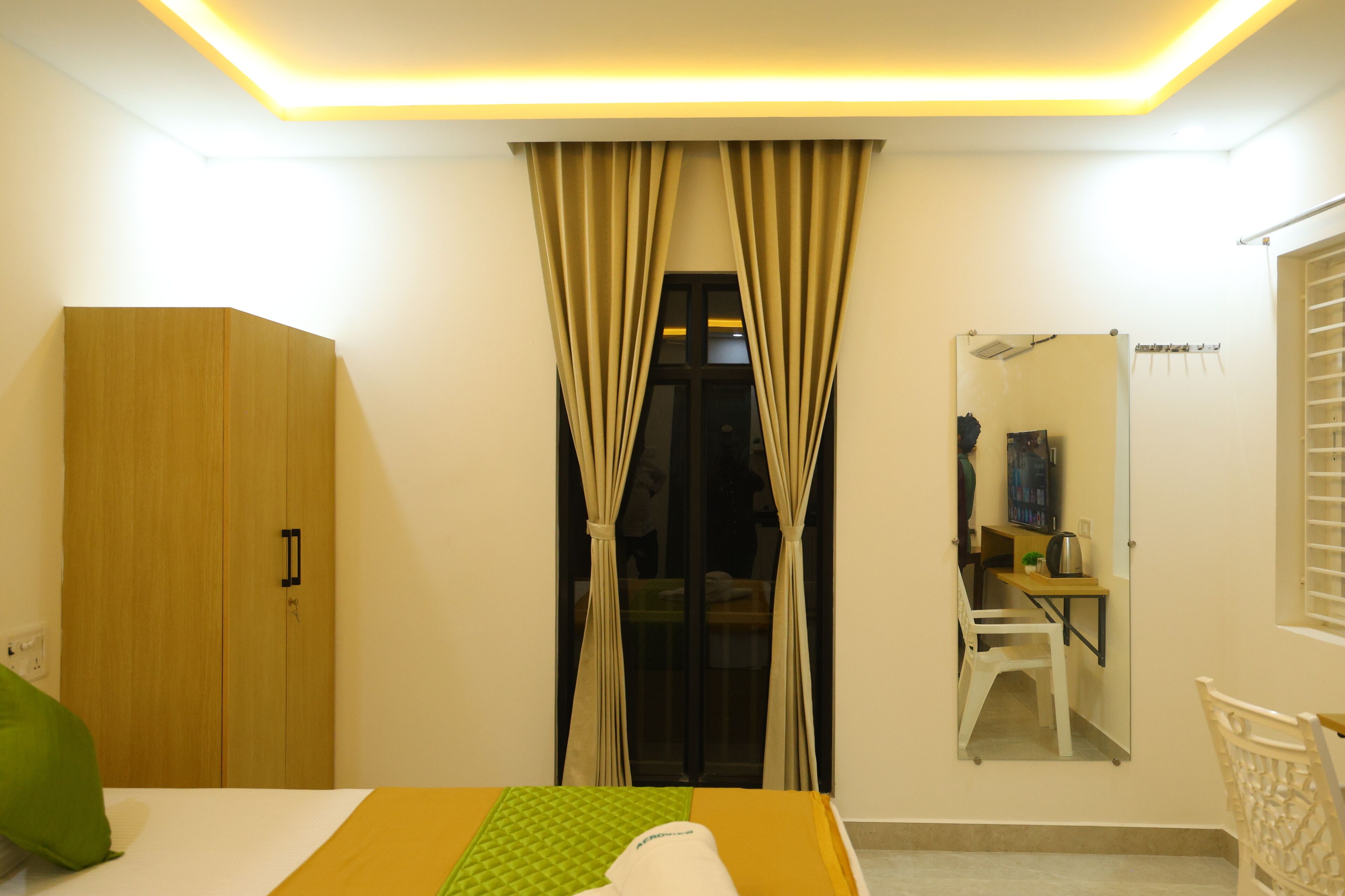 A bedroom with a bed, a wardrobe, a desk with a chair, a mirror, and a window with curtains.