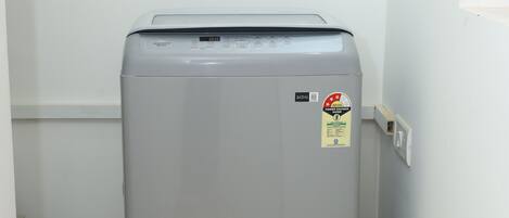 A compact, grey, wall-mounted washing machine in a narrow hallway with wooden flooring and white walls.