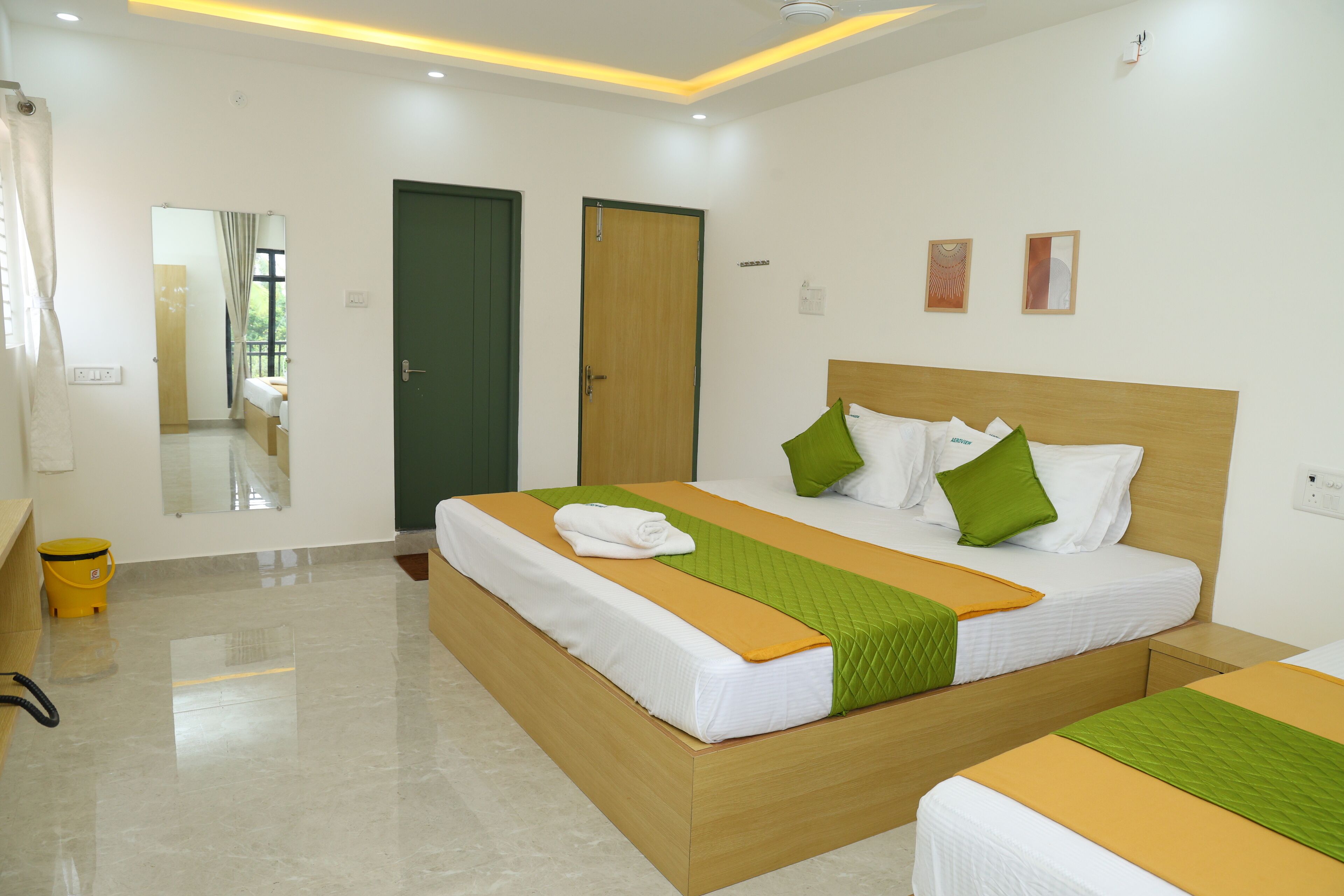 A hotel room with a large bed, a wooden headboard, and a green and orange bedspread. There is a small wooden nightstand and a yellow trash bin.