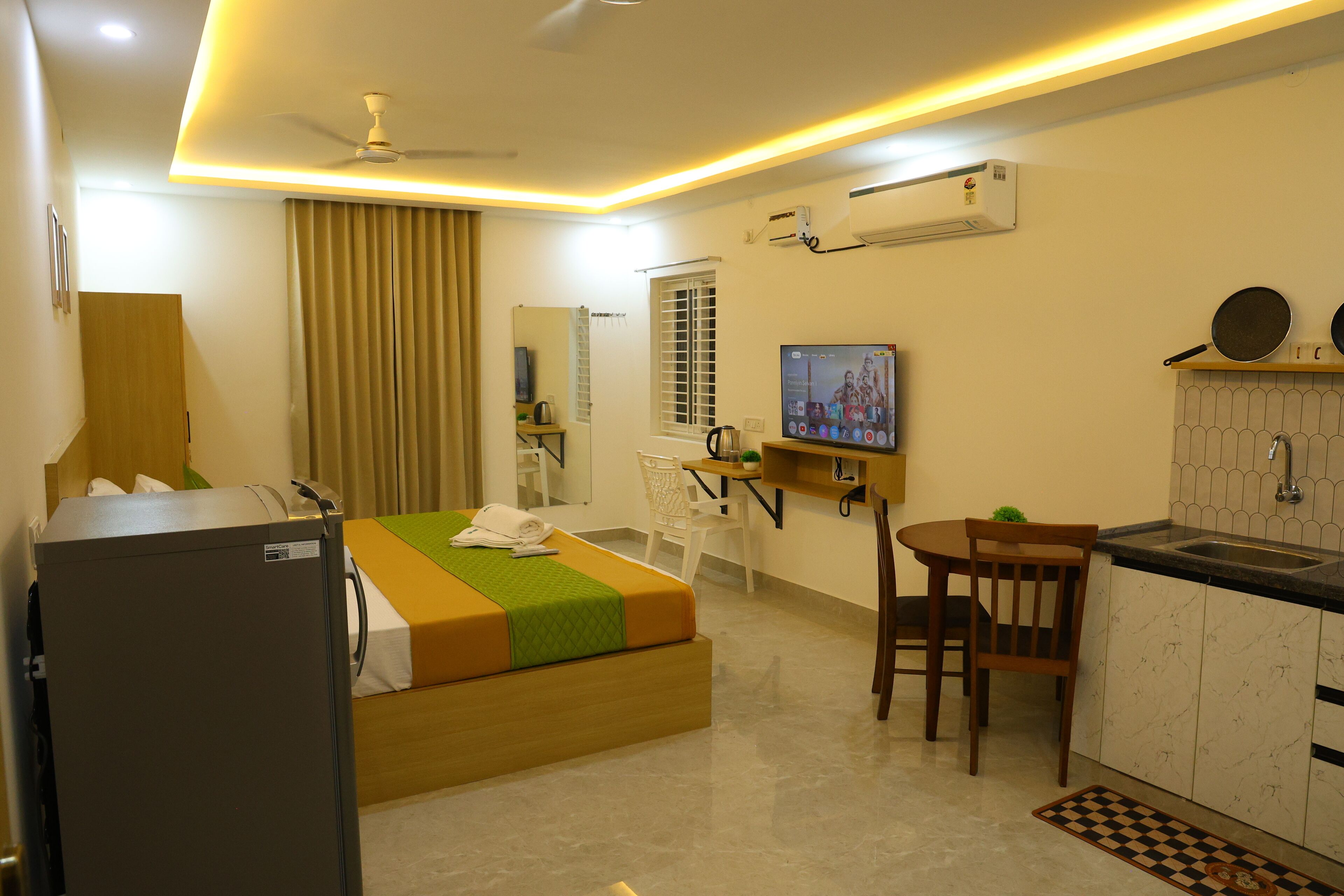 A hotel room with a bed, a desk, a chair, a television, a refrigerator, and a kitchen area.