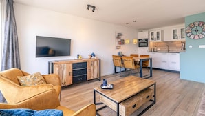A modern living room with a sofa, coffee table, and TV. A kitchen with cabinets and a clock is visible in the background. - Comfortable and Modern Furnished 4-person Apartment (De Koog)