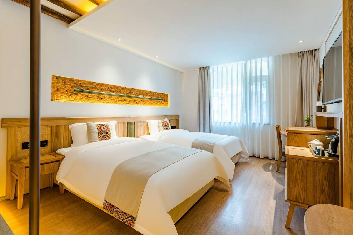 A hotel room with two beds, a wooden desk, a mirror, and a large window with curtains.