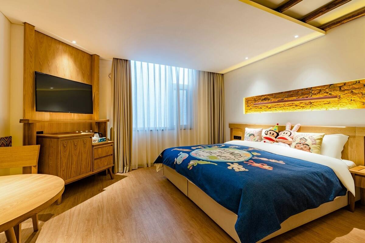 A hotel room with a large bed, a TV mounted on a wooden panel, a wooden desk, and a window with curtains.