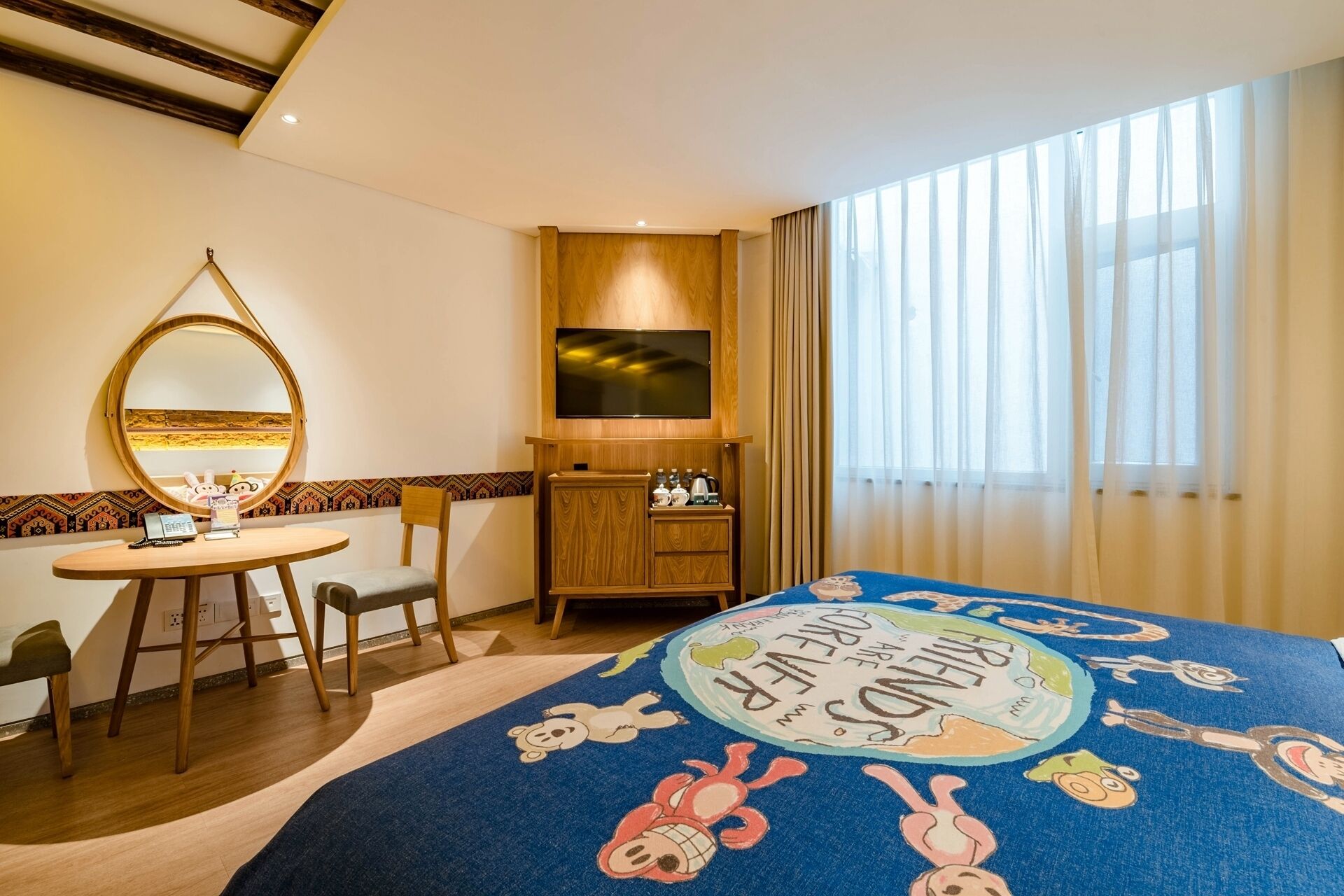 Una habitación de hotel con una cama decorada con un diseño de personaje de caricatura colorido, un escritorio de madera con una silla, un espejo, un televisor y una ventana con cortinas traslúcidas.