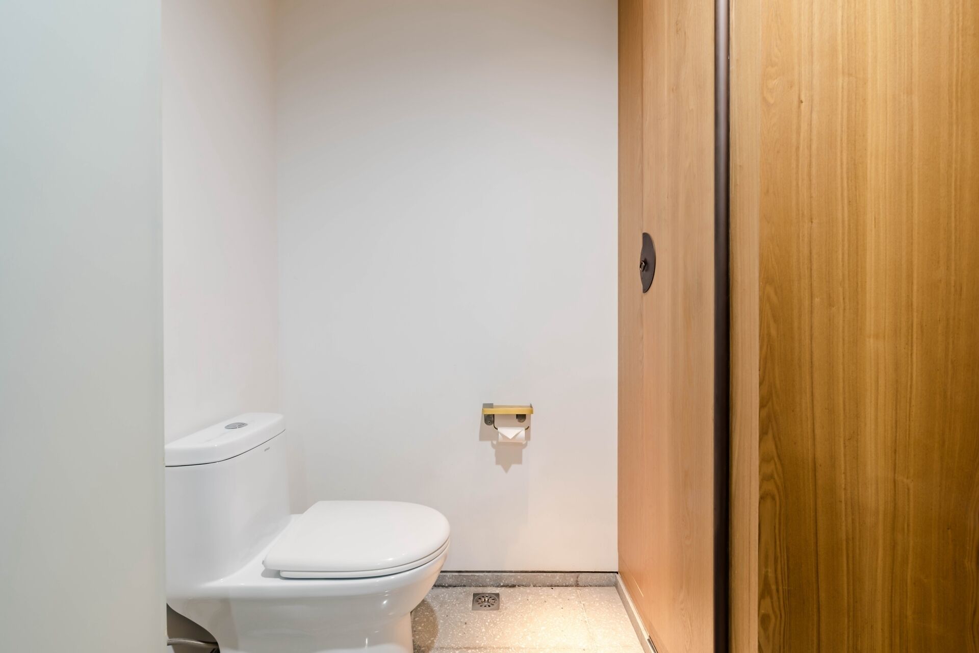 A small, narrow bathroom with a white toilet, a wooden door, and a wall-mounted light fixture.