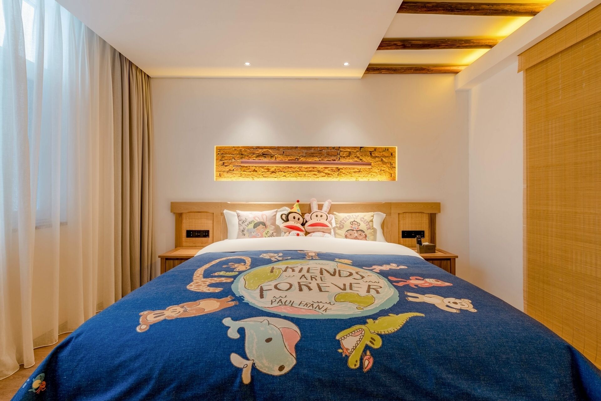 A hotel room with a bed featuring a 'Friends Are Forever' themed bedspread, a wooden headboard, a nightstand with a lamp, and a wall-mounted artwork above the bed.
