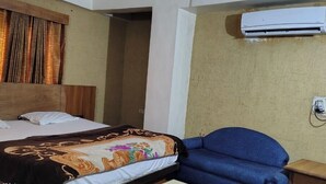 A hotel room with a bed, a blue chair, an air conditioning unit, and a power outlet. - Rajshree Hotel (Ahmedabad)