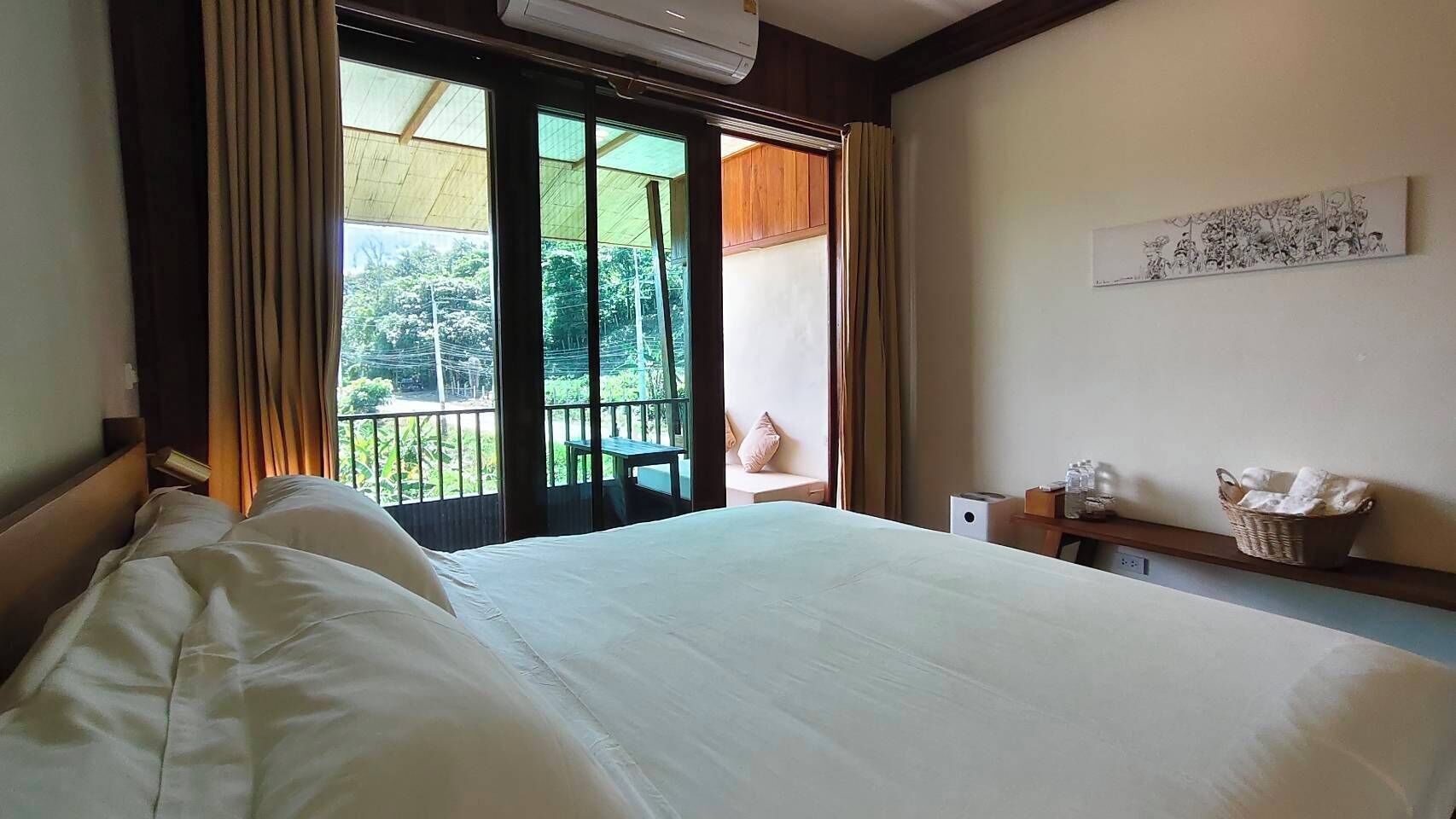 Una habitació d'hotel amb un llit gran, un balcó amb vista a la vegetació, una taula petita amb una cistella i una obra d'art penjada a la paret.
