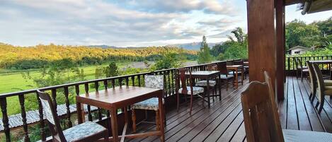 Una terraza de madera con mesas y sillas, que da a un paisaje verde con árboles y una cadena montañosa distante.