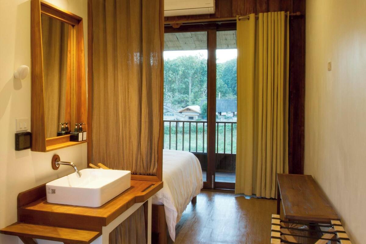 Ein Hotelzimmer mit einem hölzernen Schminktisch, einem großen Spiegel, einem weißen Waschbecken, einem Bett, einem Balkon mit Vorhängen und einer Klimaanlage.