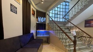 A hotel lobby with a sofa, a television, a fish tank, and a staircase with decorative railings. - Hotel Heritage (Varanasi)