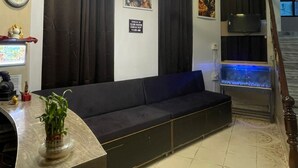 A living room with a dark sofa, a chandelier, a clock, and a sign indicating check-in times. - Hotel Heritage (Varanasi)