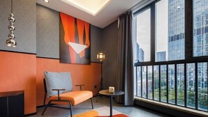 Ein modernes Hotelzimmer mit großem Fenster und Blick auf die Stadt, einer gemütlichen Sitzecke mit kariertem Sessel und minimalistischer Einrichtung, inklusive eines geometrischen Wandbildes.