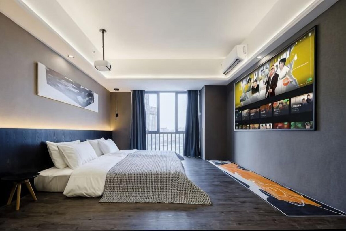 A modern hotel room with a large bed, a flat-screen TV mounted on the wall, and a large window.