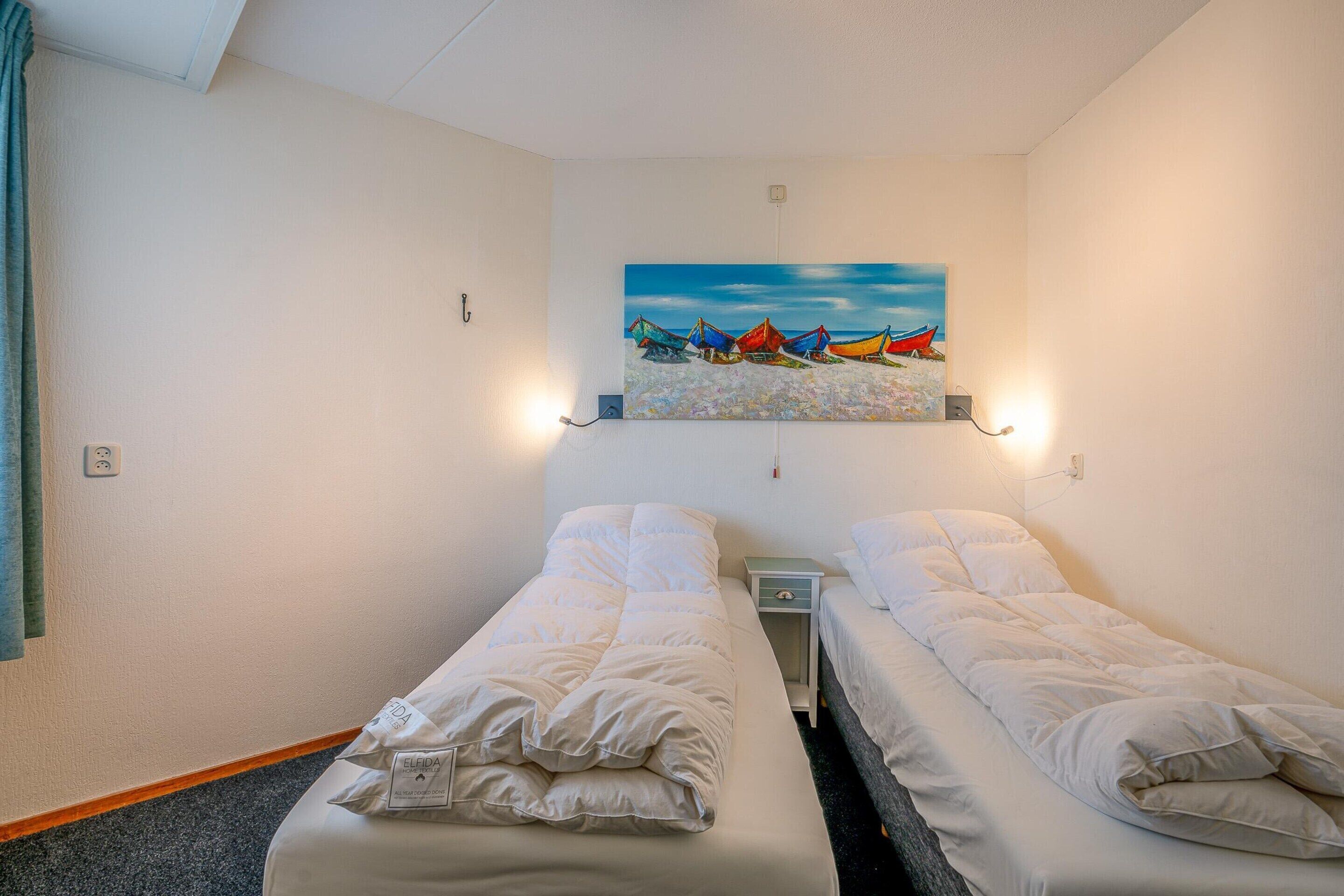 A hotel room with two single beds, a painting of boats on the wall, and a small bedside table with a lamp.